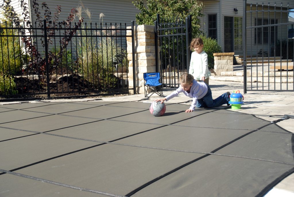 image showing kids walking on safety pool cover to retreive a ball that rolled onto it and being safely held up by the pool cover